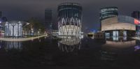 a big building by some other buildings at night time, with reflections on the water