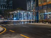 an empty city street with the lights turned on at night, and a large glass building in the background