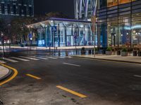 an empty city street with the lights turned on at night, and a large glass building in the background