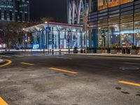 an empty city street with the lights turned on at night, and a large glass building in the background