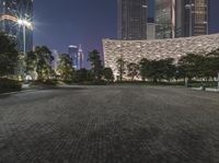 a city park with green benches and people seated on it at night in front of tall buildings