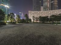 a city park with green benches and people seated on it at night in front of tall buildings