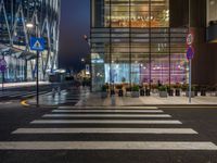 people walking and sitting on benches at a crosswalk at night in front of a glass building