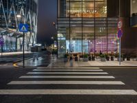 people walking and sitting on benches at a crosswalk at night in front of a glass building