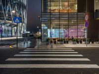 people walking and sitting on benches at a crosswalk at night in front of a glass building