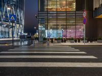 people walking and sitting on benches at a crosswalk at night in front of a glass building