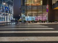 people walking and sitting on benches at a crosswalk at night in front of a glass building