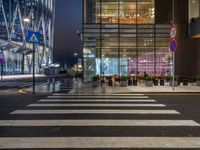 people walking and sitting on benches at a crosswalk at night in front of a glass building