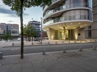 a tall circular building with glass windows on a sidewalk next to a street with a few buildings