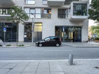 a small black car parked outside of a building on a street corner with cars in the middle