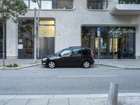 a small black car parked outside of a building on a street corner with cars in the middle