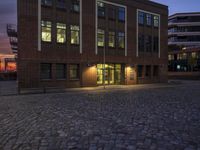 an empty bricked walkway with an illuminated building in the background with a sunset at dusk