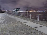 an empty brick path next to a metal fence and bridge at night with city lights in the background