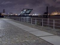 an empty brick path next to a metal fence and bridge at night with city lights in the background