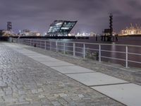 an empty brick path next to a metal fence and bridge at night with city lights in the background