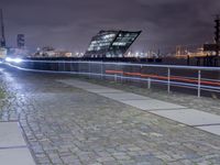 an empty brick path next to a metal fence and bridge at night with city lights in the background