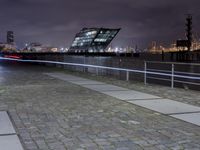 an empty brick path next to a metal fence and bridge at night with city lights in the background