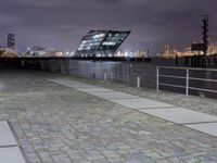 an empty brick path next to a metal fence and bridge at night with city lights in the background