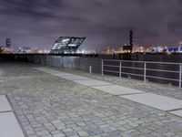 an empty brick path next to a metal fence and bridge at night with city lights in the background