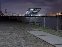 an empty brick path next to a metal fence and bridge at night with city lights in the background