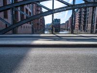 two people walking on a street with metal frame and bridge in background and buildings along river in background