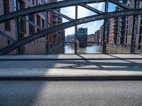 two people walking on a street with metal frame and bridge in background and buildings along river in background