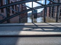 two people walking on a street with metal frame and bridge in background and buildings along river in background