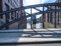 two people walking on a street with metal frame and bridge in background and buildings along river in background