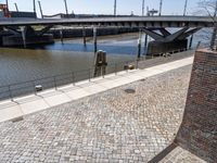 the view of the bridge from an area with brick walkways and water in background