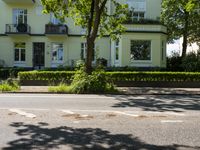 an empty street and the corner of a two - story white house with green hedges on both sides