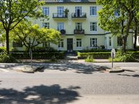 an empty street and the corner of a two - story white house with green hedges on both sides