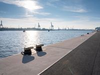 the two wooden benches are overlooking the large body of water that has many piers and cranes in the background