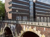 large brick building with tall glass and metal buildings behind it, near bridge over river