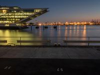 a photo of some benches in front of a body of water at night with the lights reflecting off the glass