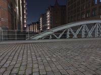 a brick walkway with a white bridge on it at night, surrounded by buildings and tall buildings