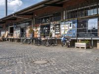 people with bicycles are walking on the cobblestones at an outdoor area with some tables and bars