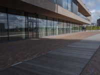 the walkway is lined with small bricks and wooden planks, along the side of a building with large windows