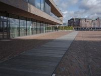 the walkway is lined with small bricks and wooden planks, along the side of a building with large windows