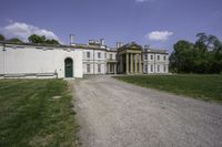 a graveled path leads to the front of a large white mansion that is next to a field