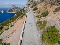 the road is surrounded by rocks and trees and near the ocean with mountains and water in background