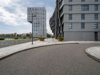 two buildings in an open city with a paved driveway and sidewalk and small planters with trees to the side