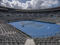 a tennis court has empty seats and a blue floor with a sky background and gray concrete steps leading to the upper level