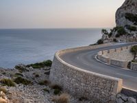 a car driving along a curved road next to the water and mountains with a light house in the distance