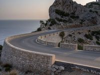 a car driving along a curved road next to the water and mountains with a light house in the distance