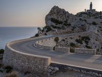 a car driving along a curved road next to the water and mountains with a light house in the distance