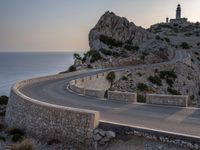 a car driving along a curved road next to the water and mountains with a light house in the distance