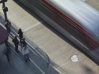 people stand in front of a red and silver train going past a station on the road