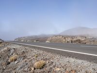 a road near the mountain side in the foggy sun day, and on the opposite bank are hills