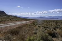 dirt road through dry brush and grasses near mountains with clouds above them and distant sky with sparse vegetation