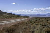 dirt road through dry brush and grasses near mountains with clouds above them and distant sky with sparse vegetation